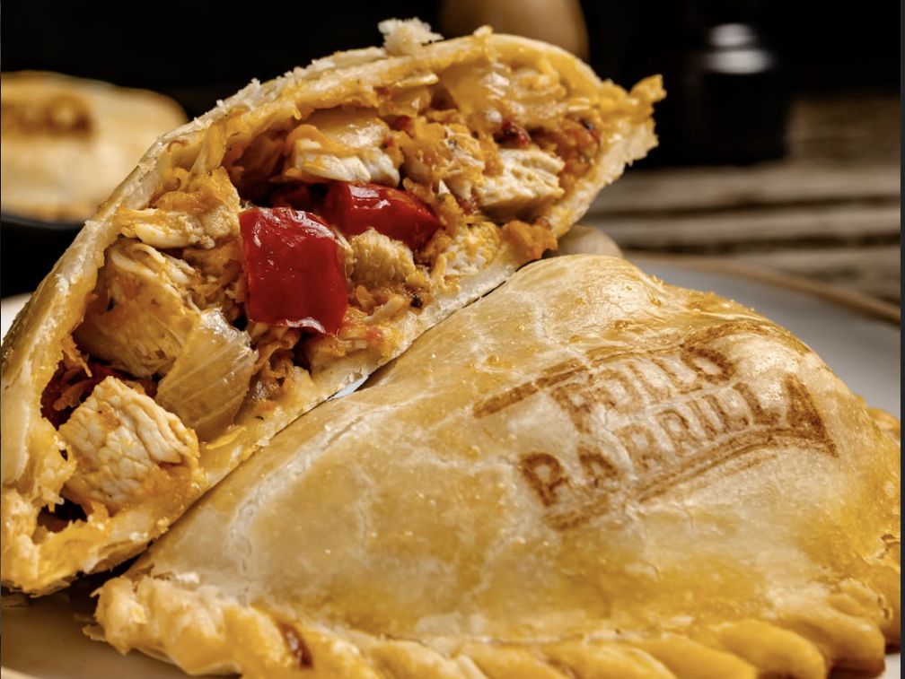 Empanada de pollo a la parrilla partida al medio, con pollo y morrones asados a la vista, junto a otra entera con el sabor marcado en la masa.